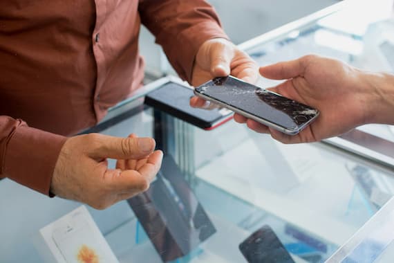A customer hands over a broken smartphone with a smashed screen to a salesperson behind a glass display case